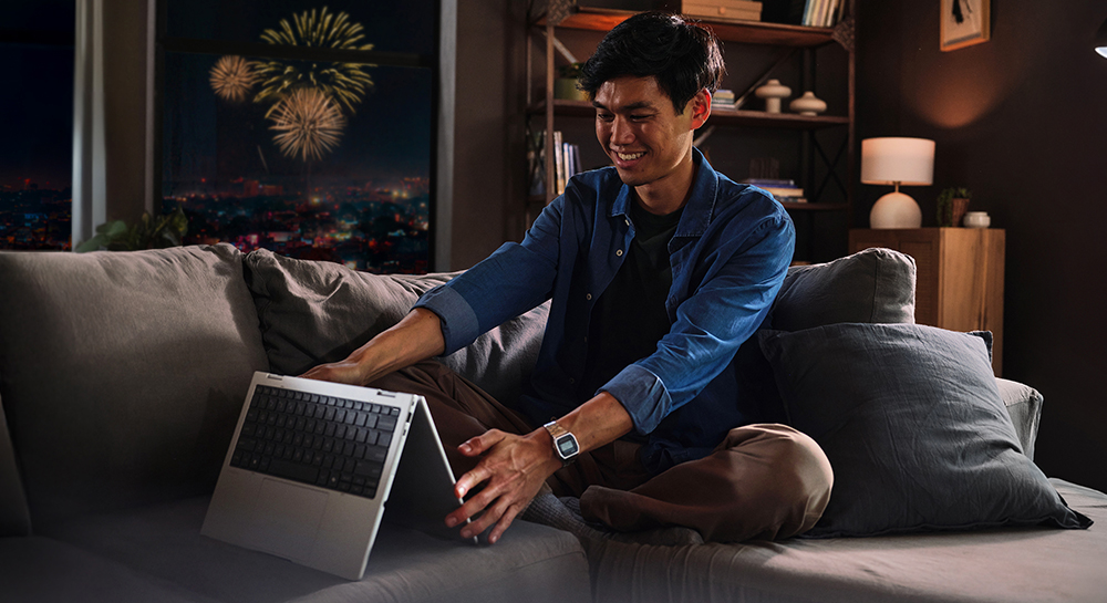 Imagen de un hombre sentado en la sala de su apartamento con una notebook x360 HP. Ambiente nocturno con juegos artificiales vistos desde la ventana.